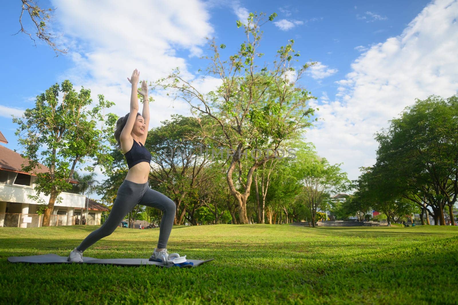 Fit asian woman practicing Warrior one pose on a mat in the park.