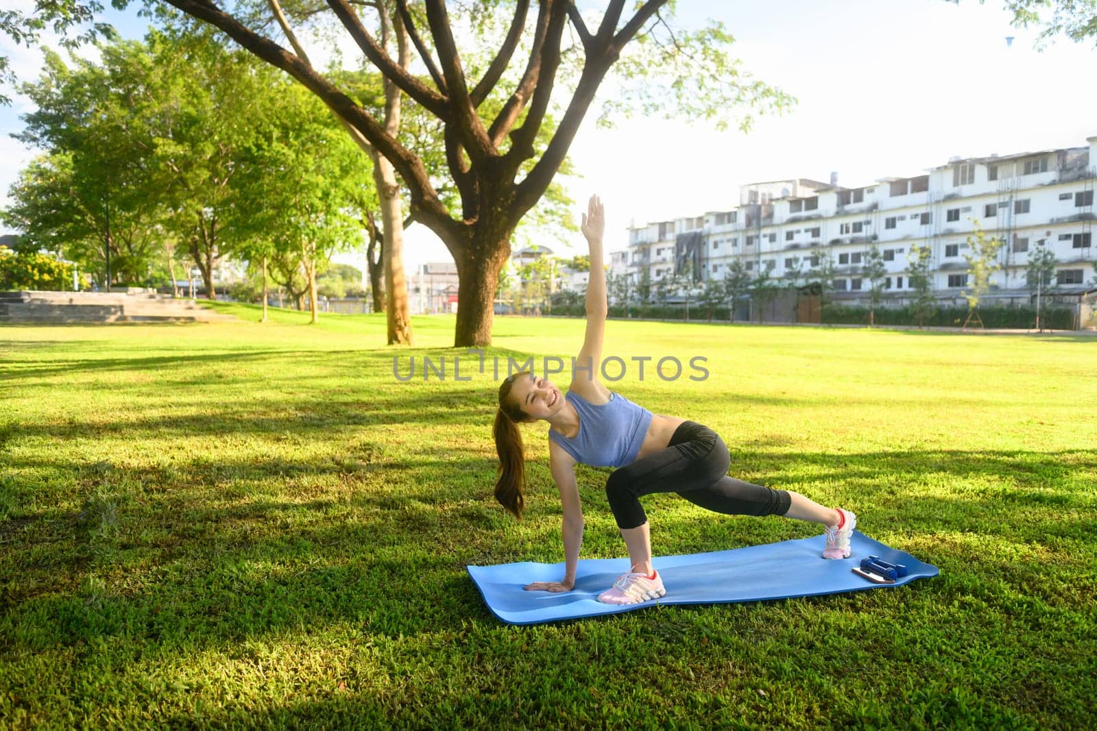 Fit asian woman practicing yoga outdoors stretching with one arm raised on mat.