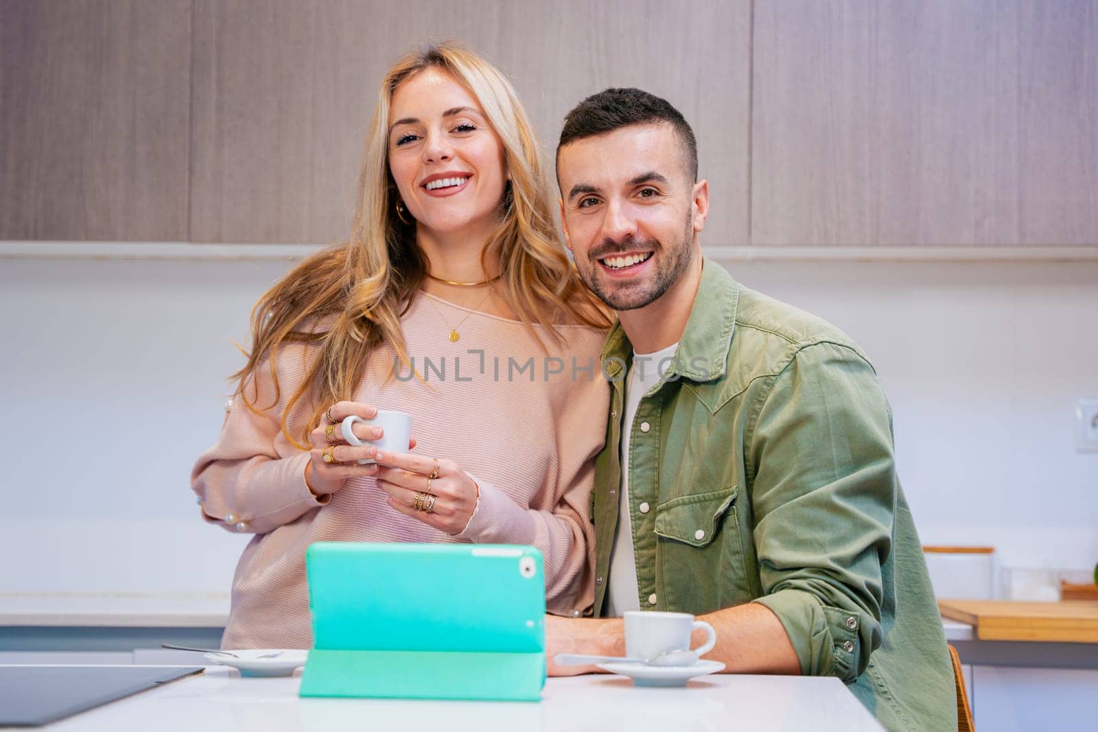 Happy couple sitting together in a modern kitchen, smiling while enjoying coffee and using a green tablet, representing connection,domestic lifestyle, technology,and relaxed morning moments at home.
