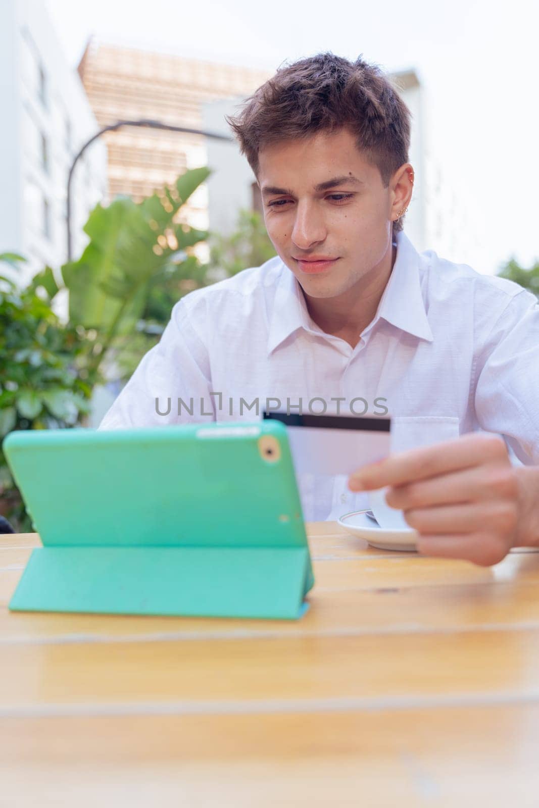 Smiling young man holding credit card while using tablet at an outdoor cafe. Concept of online shopping, e-commerce, and digital payment in a modern, casual, and urban setting.Vertical