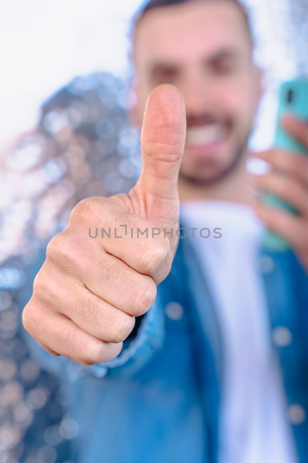 Happy young man in denim shirt holding a smartphone and giving a thumbs up with a big smile, standing in front of a modern reflective wall, representing approval, success, and digital connection. Vertical