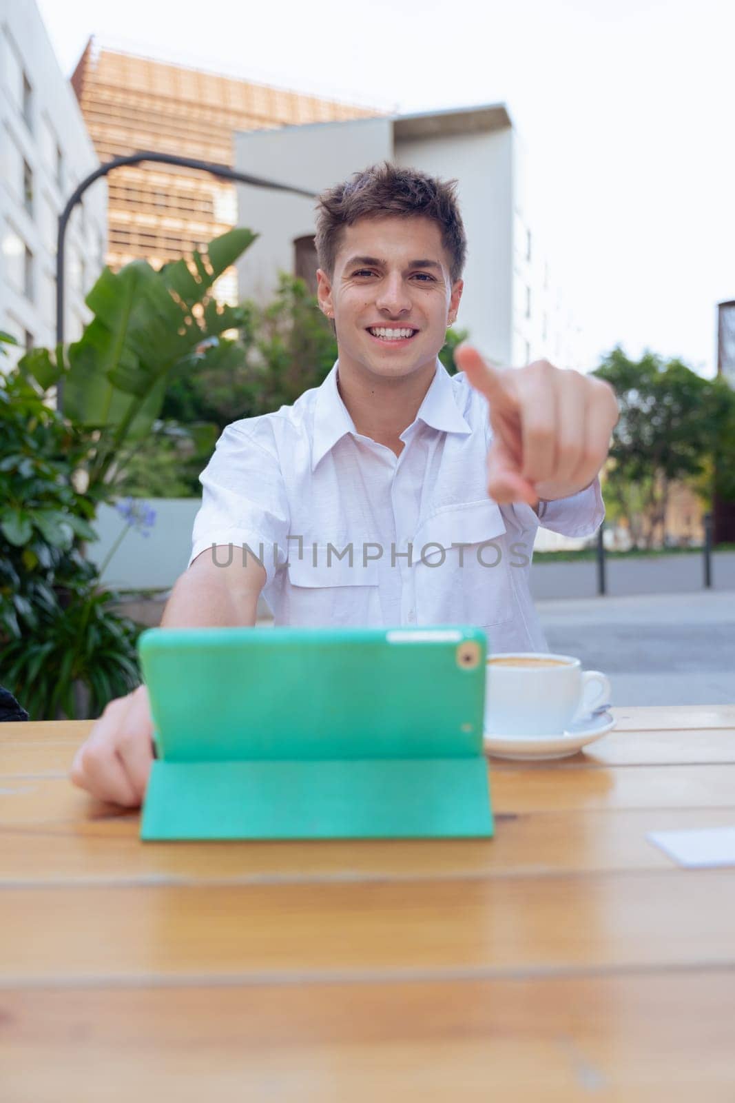 Vertical photo of a young man smiling and pointing at the camera while sitting outdoors with a tablet and cup of coffee on a wooden table, symbolizing communication, positivity,and digital lifestyle