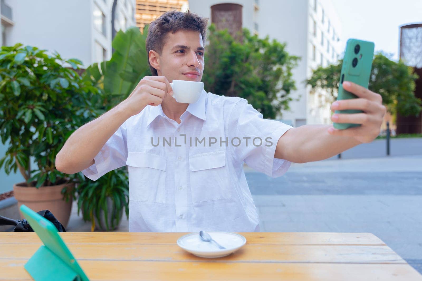 Young man in white shirt sitting at an outdoor table with a tablet, smiling while holding a cup of coffee and taking a selfie, representing lifestyle, connection, technology, and relaxed moments.