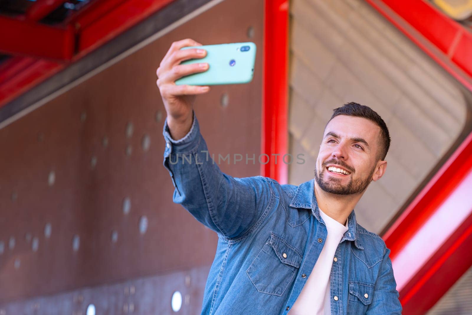 Young man smiling while taking a selfie with his smartphone in an urban environment. Casual and modern moment with red architectural elements in the background, showing confidence and joy.