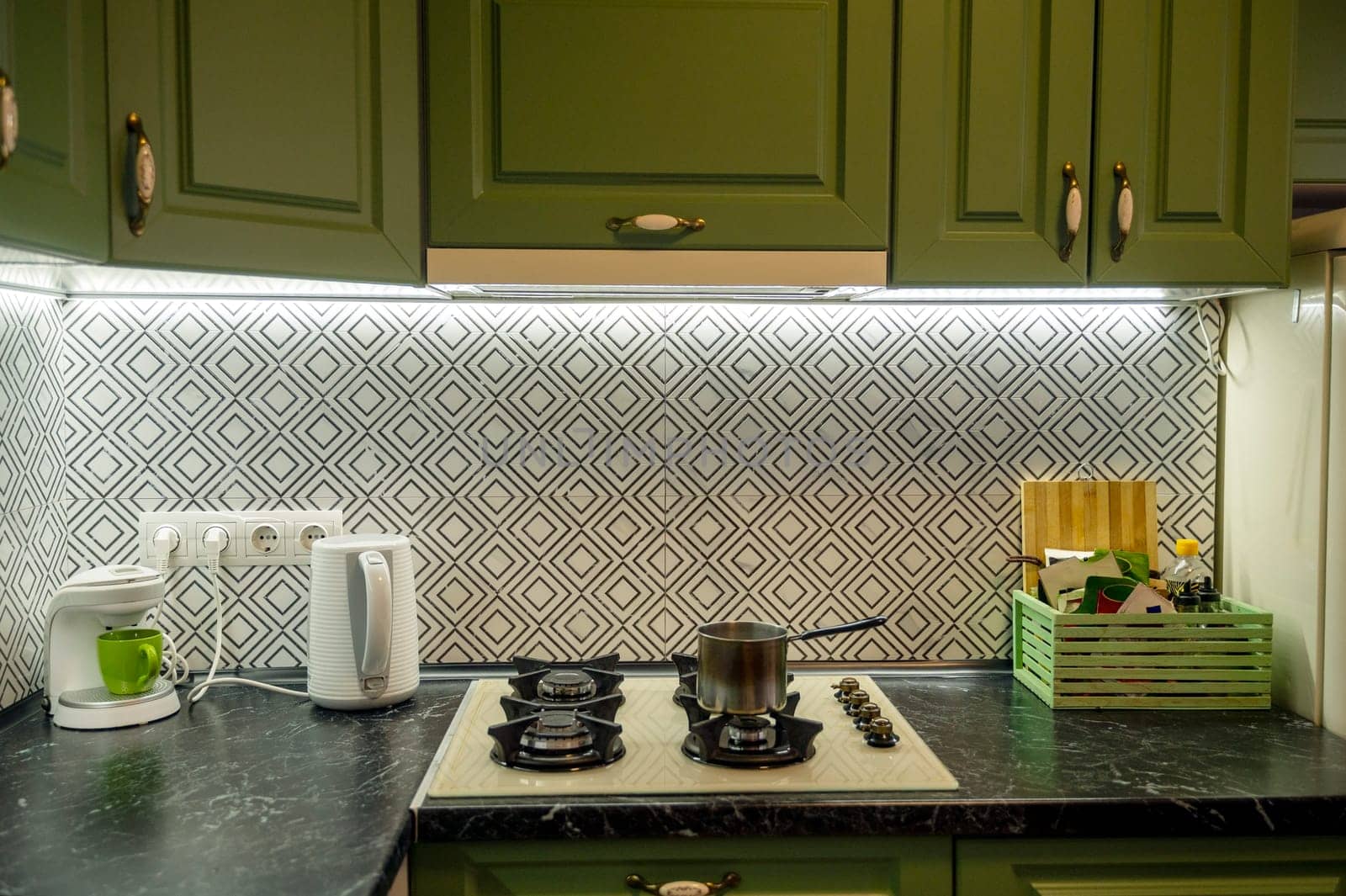 Detail of the kitchen with countertops, gas stove, green cabinets, kettle and coffee maker. On the stove there is a saucepan, a drawer for spices. No brands or logos.