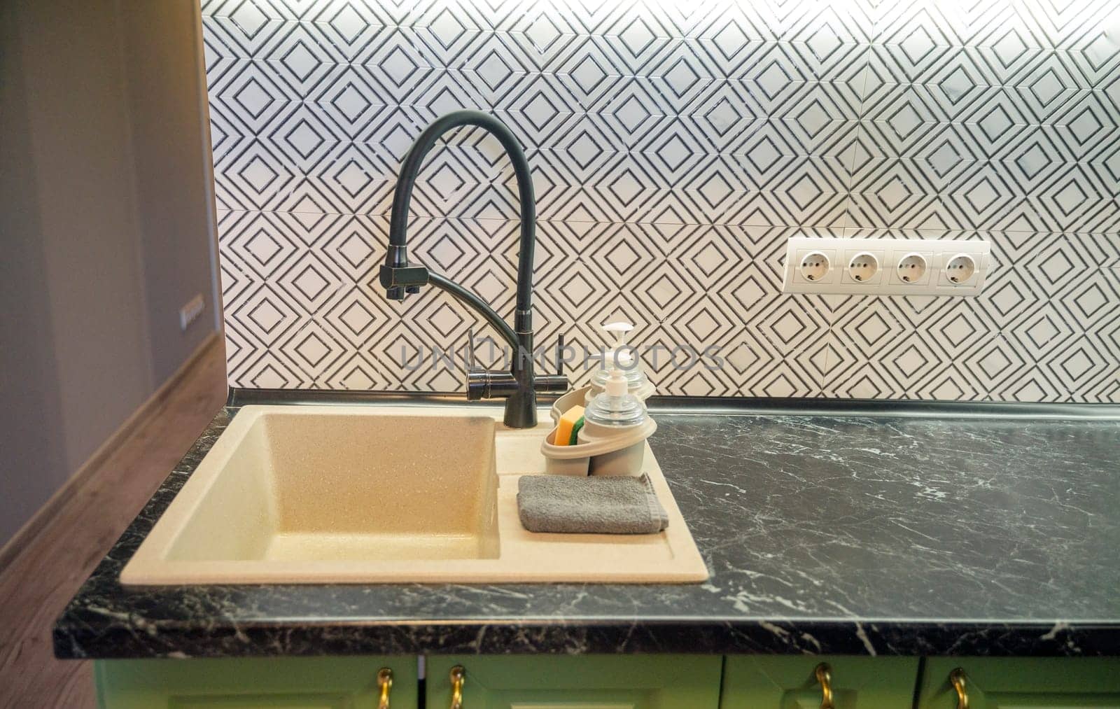Close-up of a light beige kitchen sink with a modern black faucet, dark green marble countertop, and a black and white tile backsplash. Dishwashing bottles are nearby.