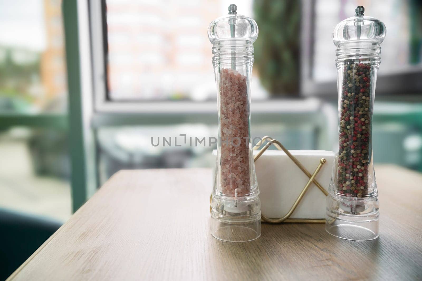 Two elegant glass salt and pepper shakers sit gracefully on a wooden dining table, along with paper napkins. Salt and pepper mills on a wooden table and paper napkins. Pink salt, coarse grind.