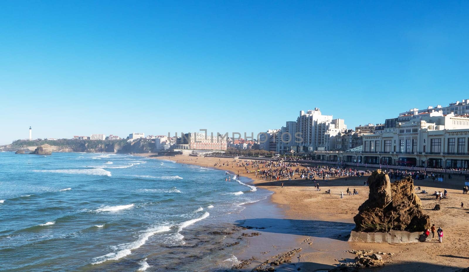 A beach with a large rock on it and a lot of people. The beach is crowded and the water is calm, Biarritz, France