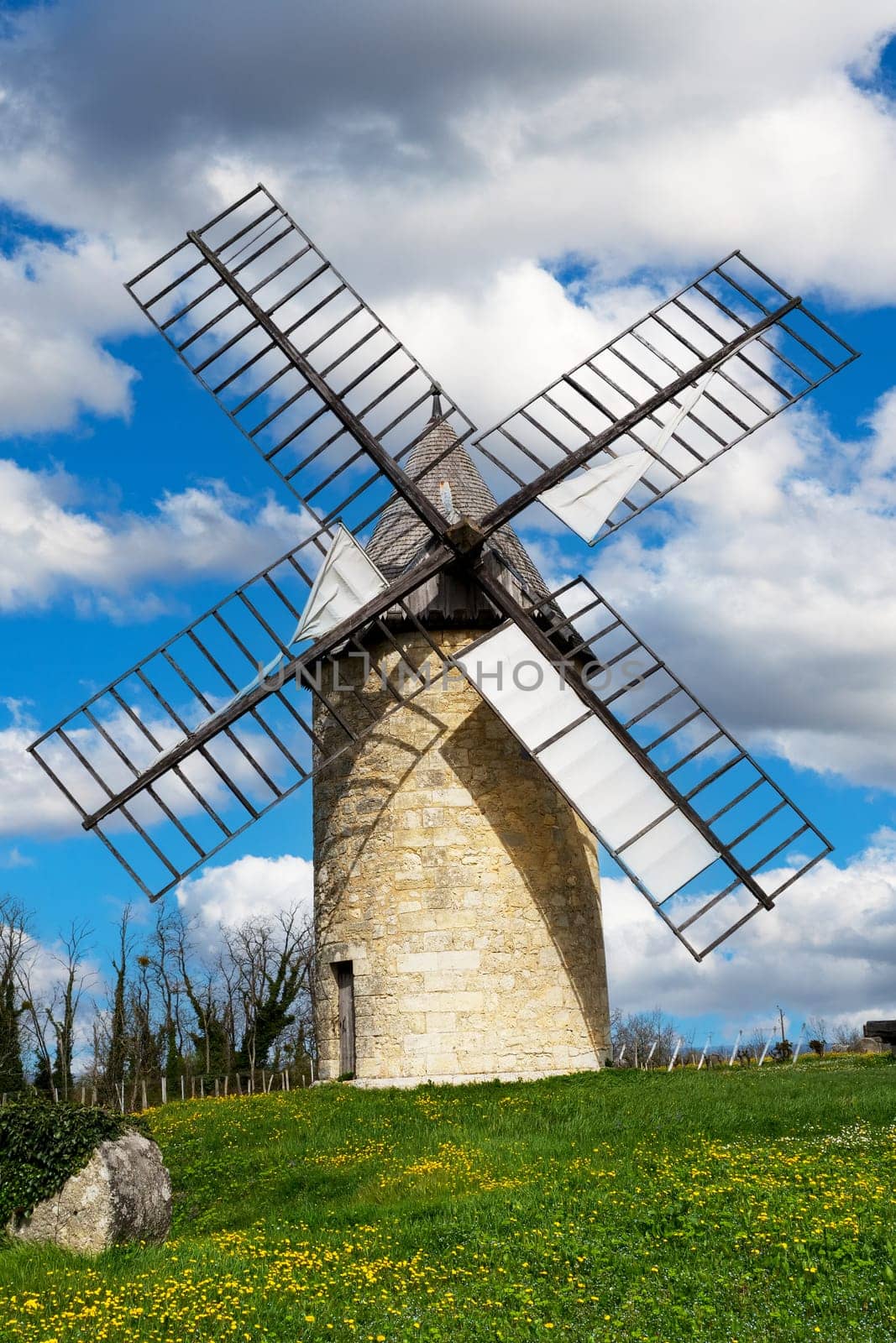 A windmill is on a hill with a field of yellow flowers. The windmill is tall and has a lot of metal bars. The sky is blue with some clouds