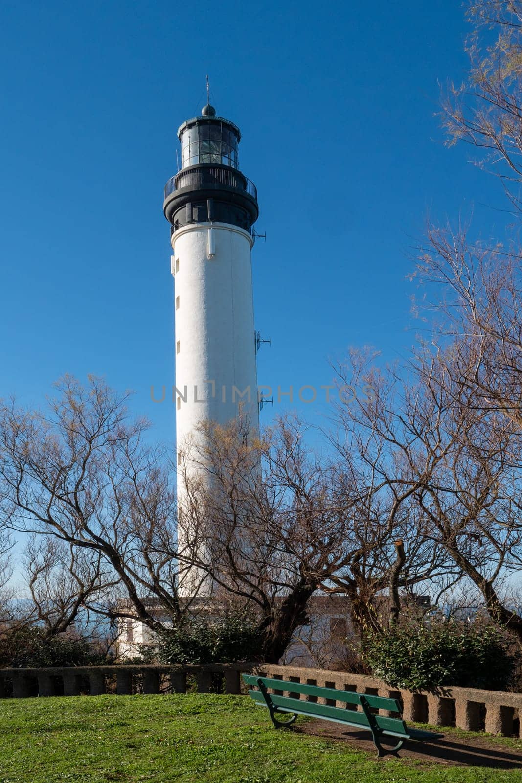 A tall white lighthouse is in the foreground of a green park. The park is empty and the sky is clear, Biarritz, France