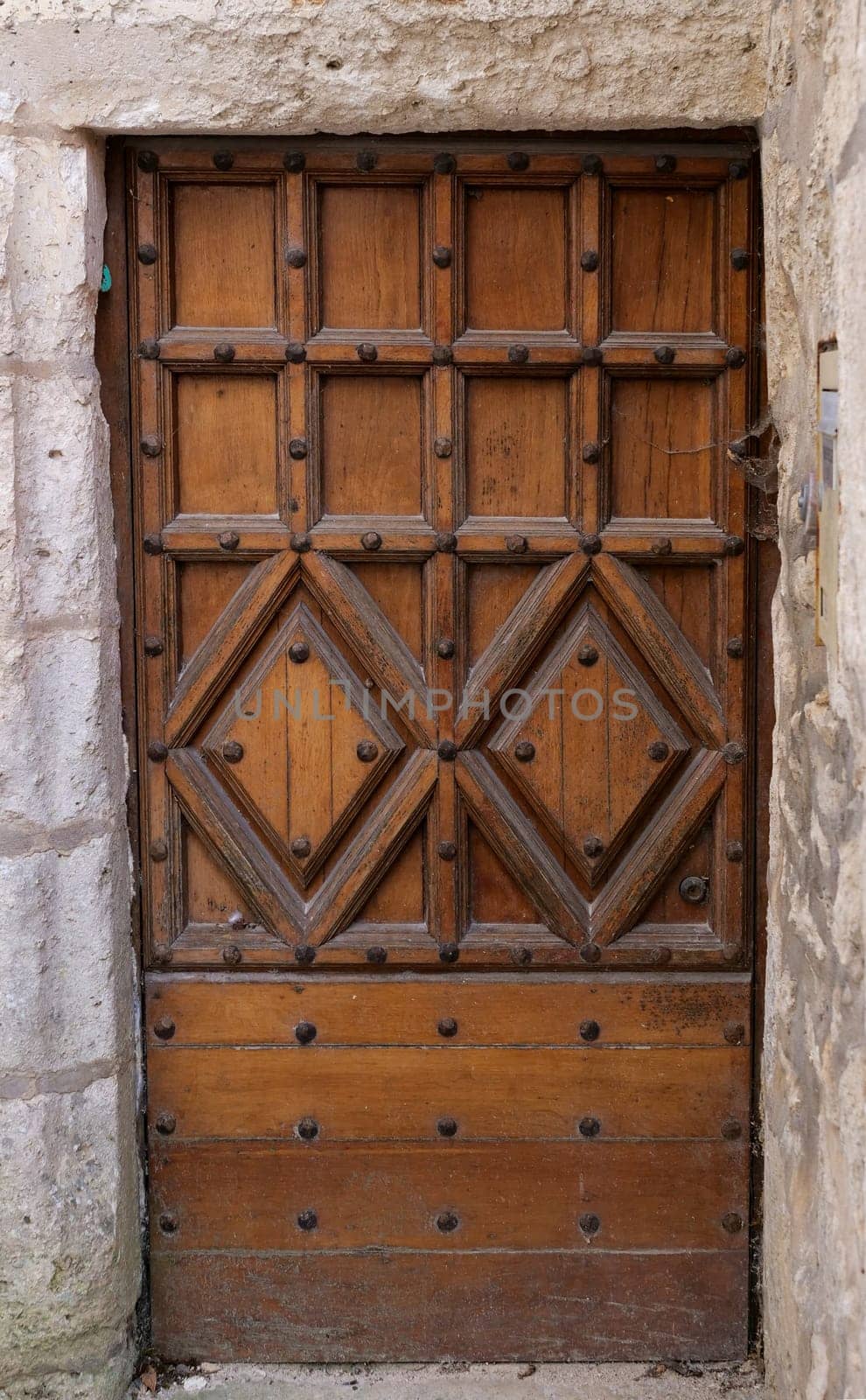 A wooden door with a stone frame and a stone wall behind it. The door has a diamond pattern on it and is covered in small holes