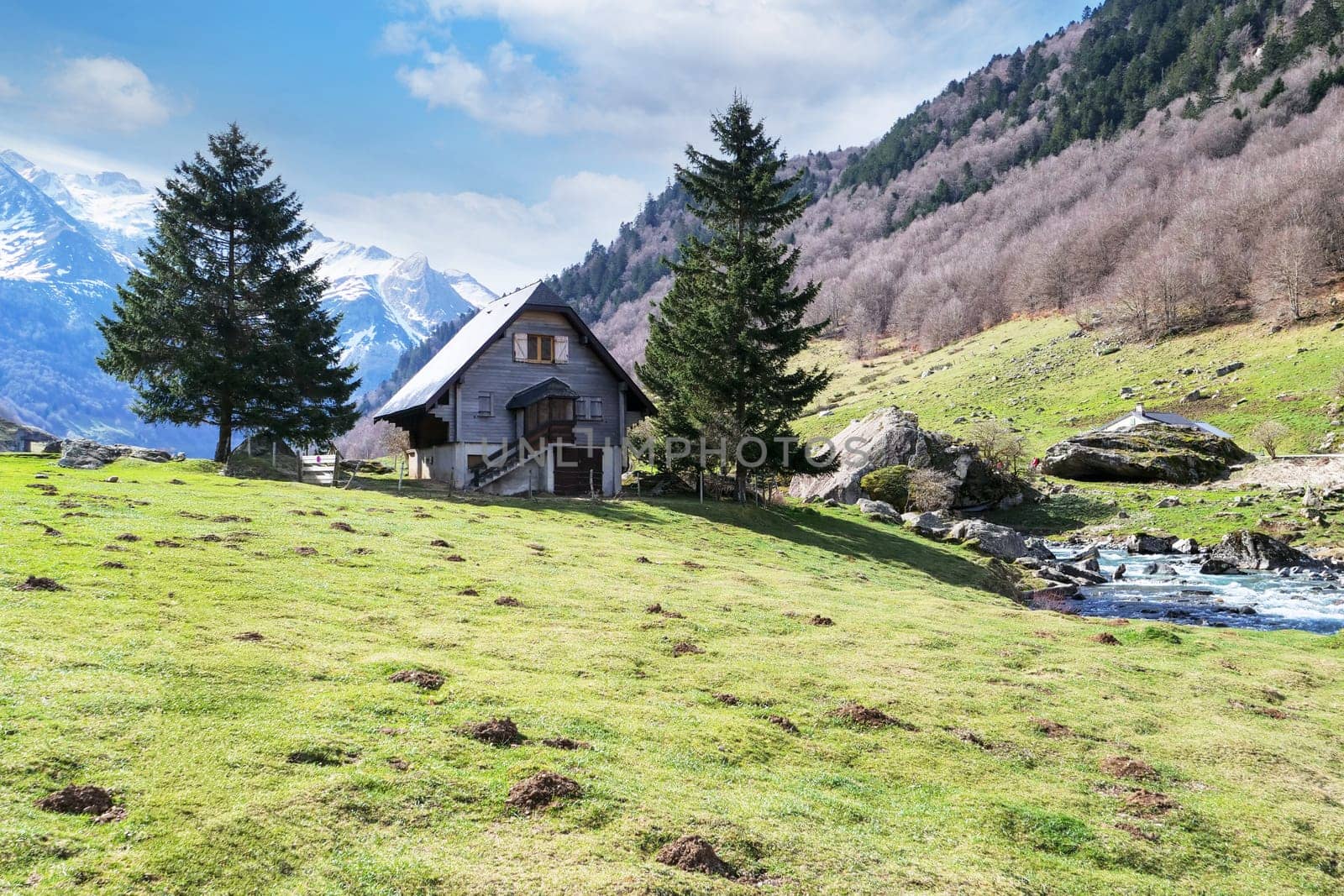A small house sits in a grassy field with a stream running nearby. The house is surrounded by trees and the mountains in the background. The scene is peaceful and serene