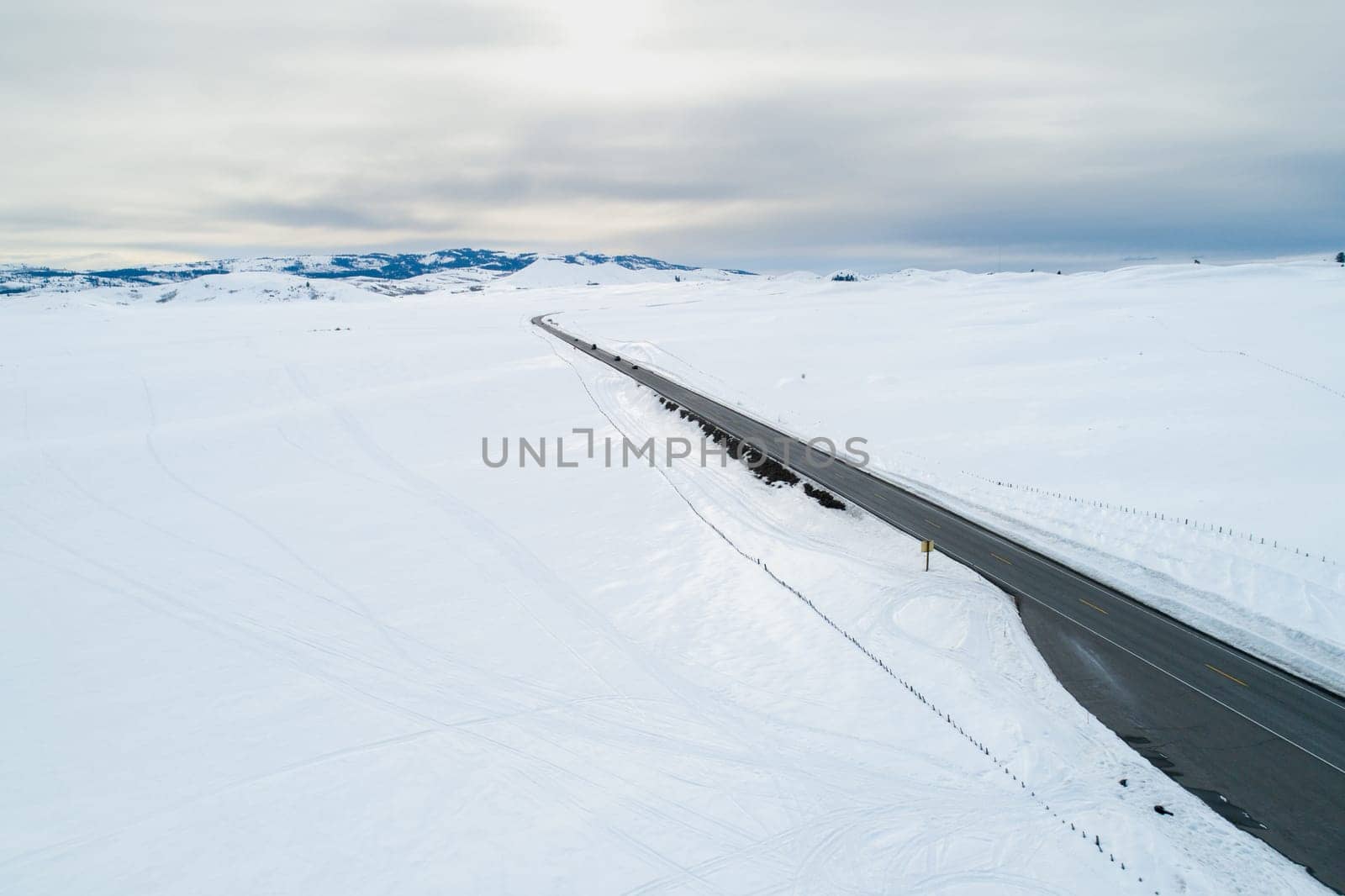 A road in the snow with a yellow sign on the side. The road is empty and the sky is cloudy