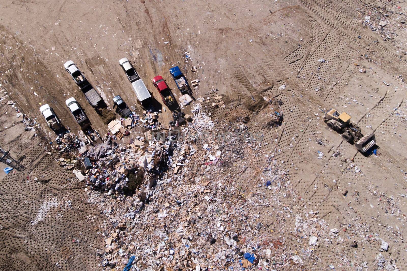 A pile of trash and a few trucks are in the background. The trucks are parked in a line, and the pile of trash is in the foreground. Scene is one of waste and pollution, with the trucks