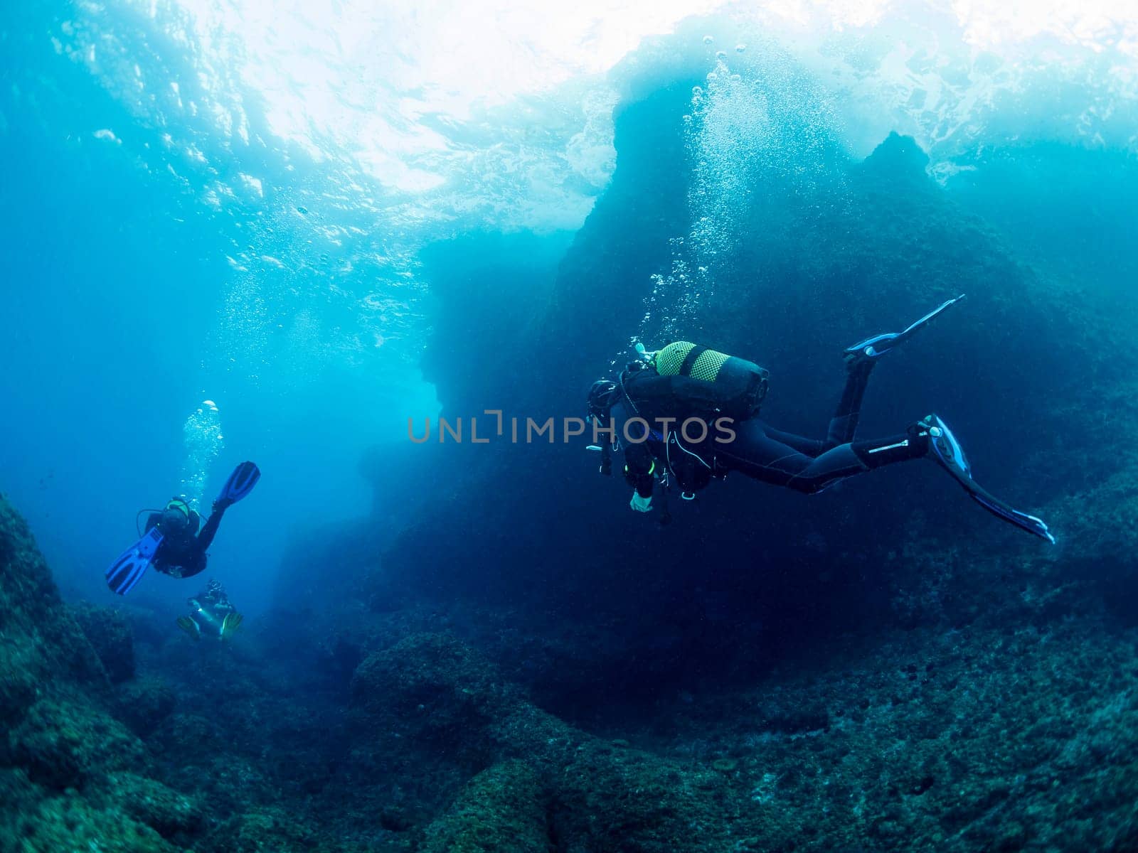 group of three divers diving through the crystal clear water with the sun reflecting on the sea surface, concept of recreational scuba diving and underwater adventure