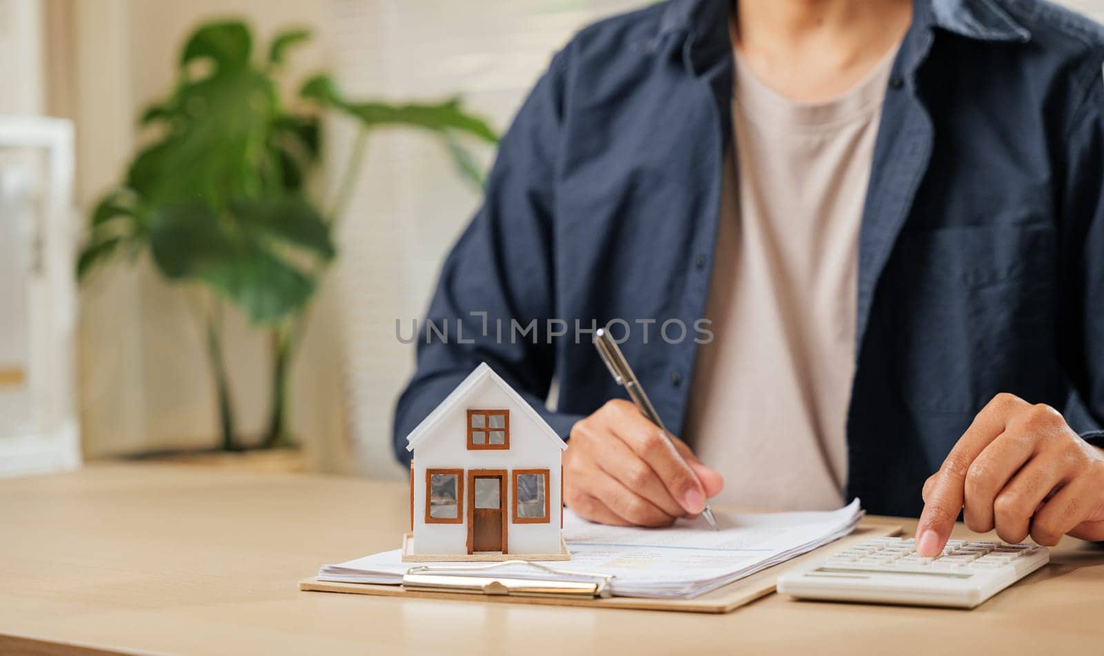 A man writes a signature on house insurance forms, calculating property tax. Using a pen and model house, he plans price, rent, and payments for property investments and contracts.