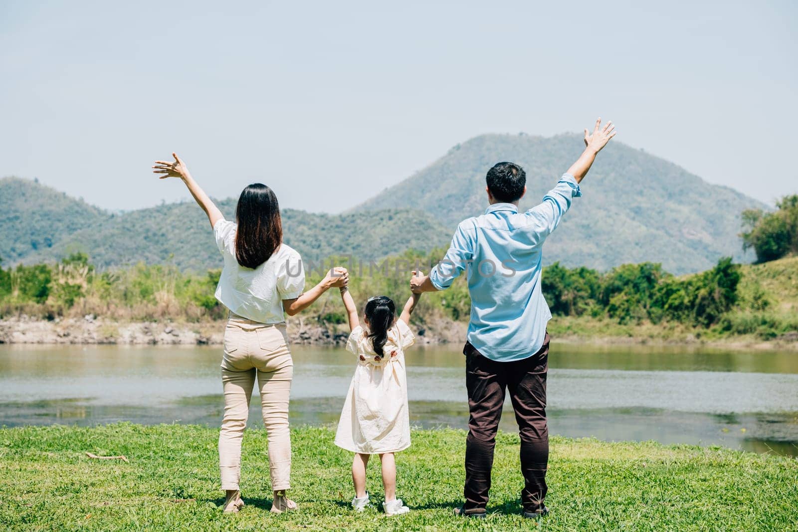 Asian family father mother and daughter stand front lake with raised hands sunny day in park, lifestyle parents and child kid enjoying outdoors together in nature on summer travel, Family relaxation