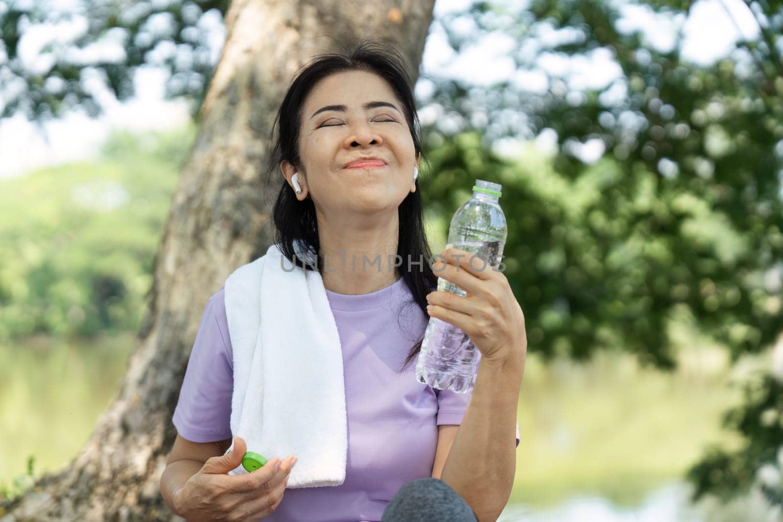 A happy woman sits by a serene lake, savoring hydration after exercise, embodying wellness and self-care.
