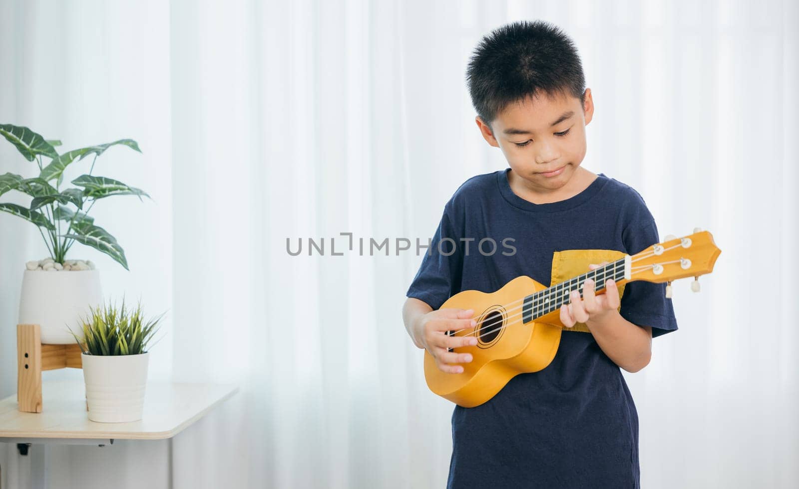 Cute Asian school boy in a blue shirt enjoying playing a yellow ukulele in living room. The soft light from the white curtain creates a warm, inviting atmosphere as he strums his instrument happily.