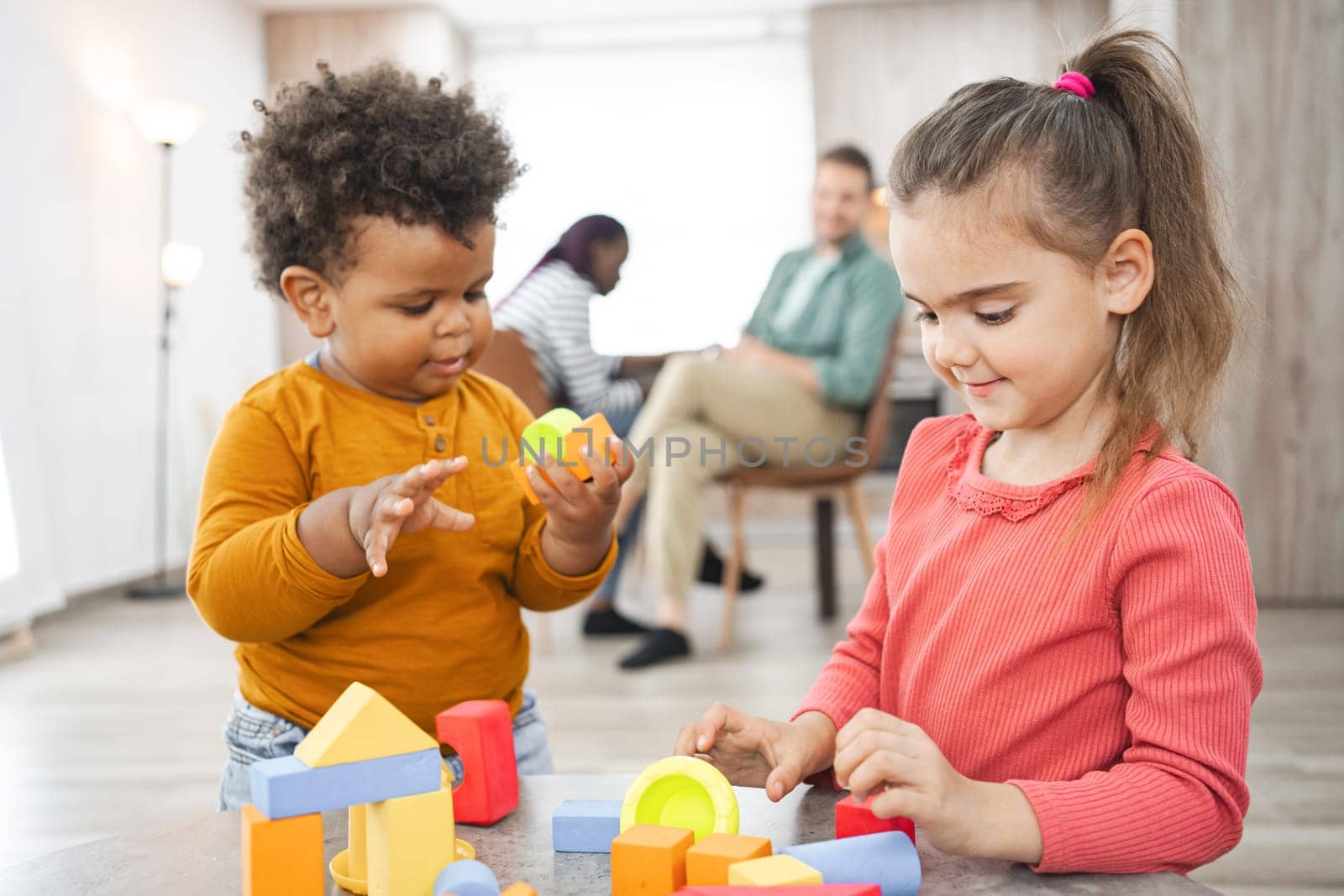 Two children play with colorful blocks while adults relax in background.