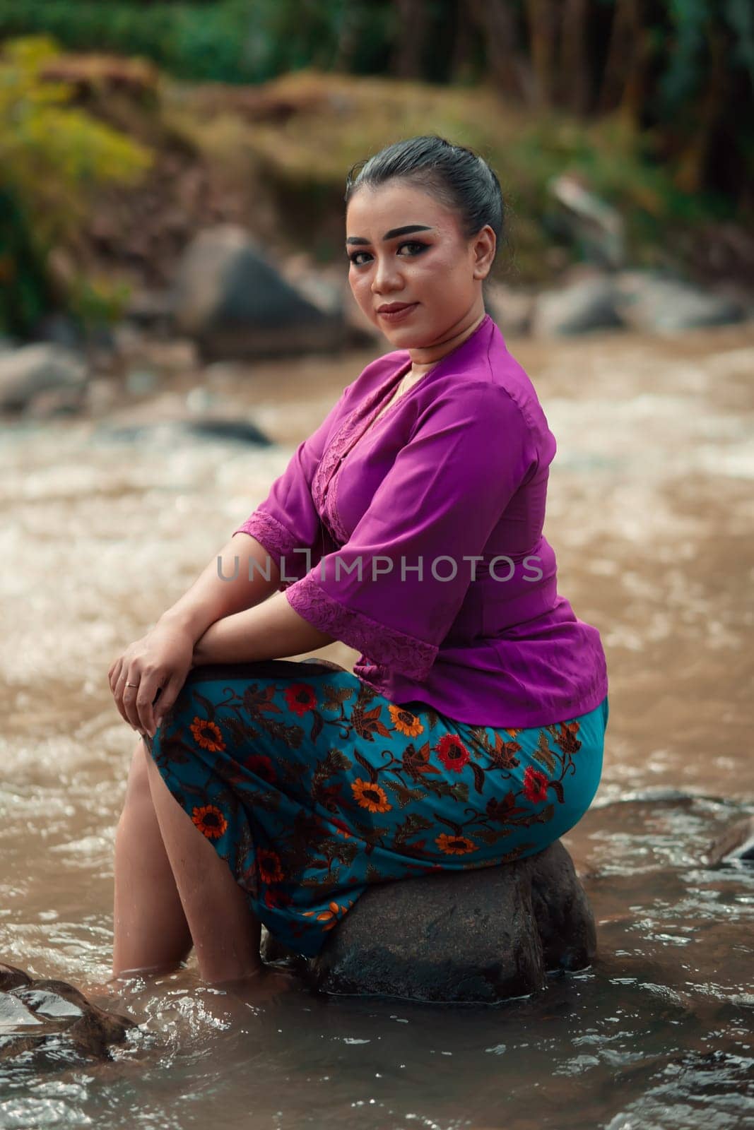 Beautiful Asian woman sitting on the rock near the river with a smiling face while wearing a purple dress inside the village