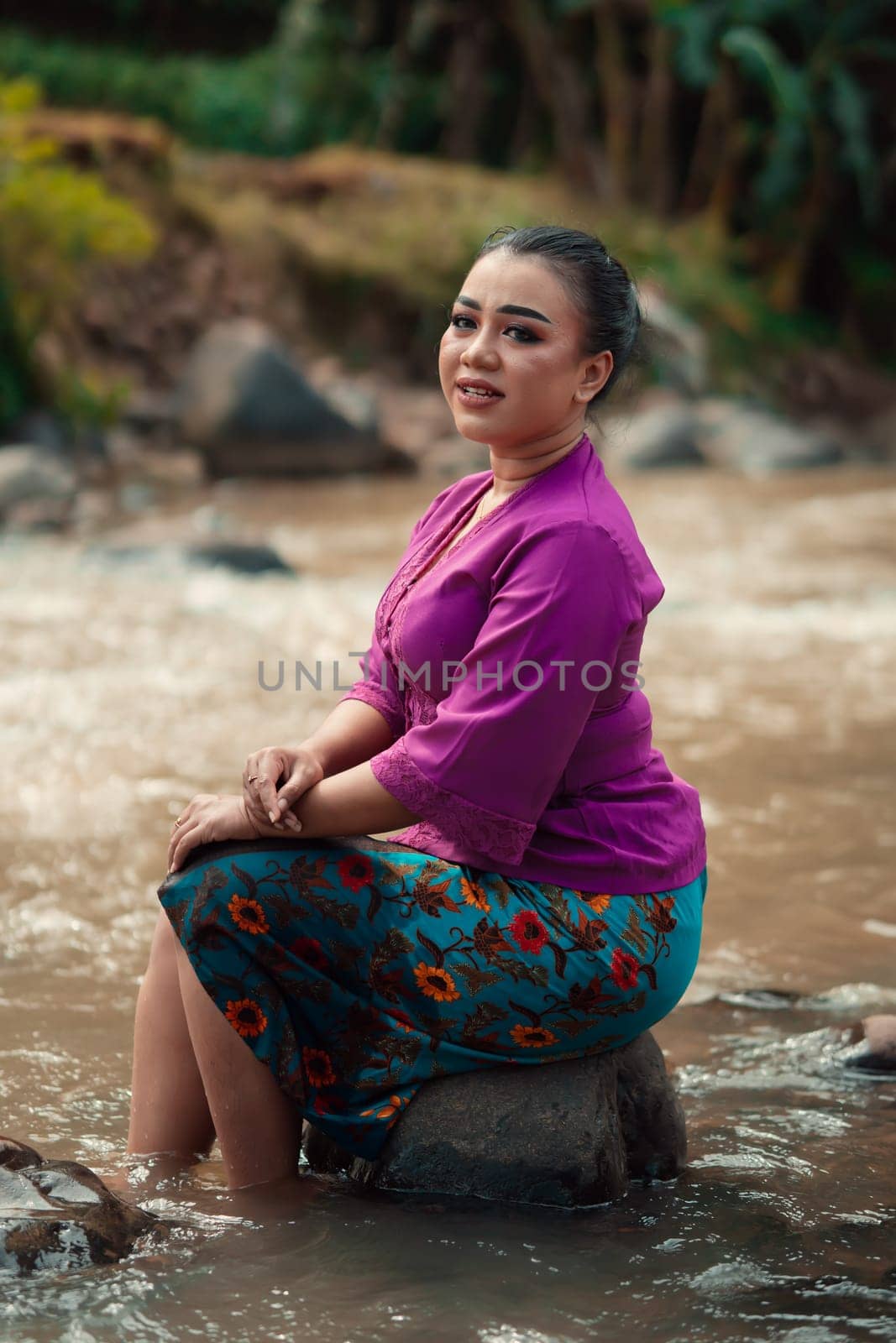 Indonesian woman with purple clothes and makeup on her face sitting near the river with brown color water while visiting the river for vacation