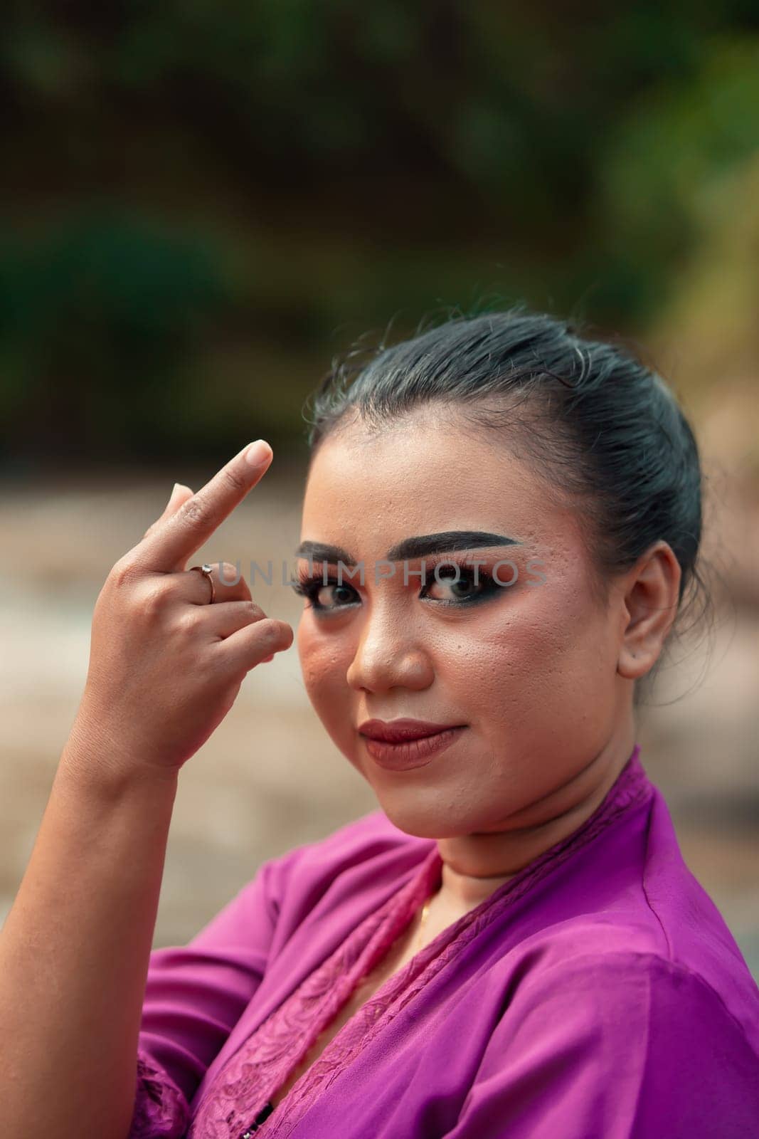 Beautiful Javanese woman with makeup and purple dress posing with her hand while sitting on the rock near the river inside the village