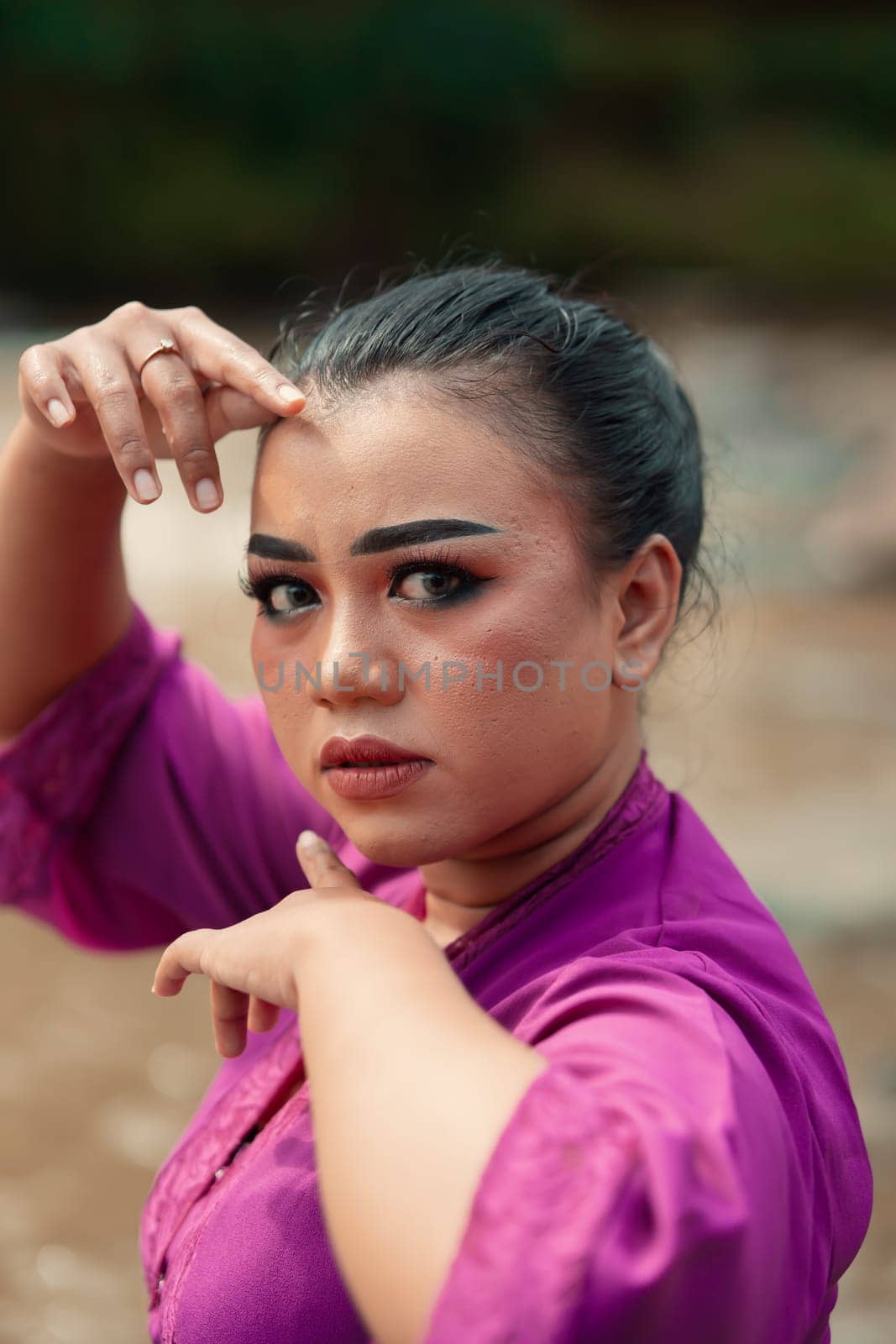 Beautiful Javanese woman with makeup and purple dress posing with her hand while sitting on the rock near the river inside the village