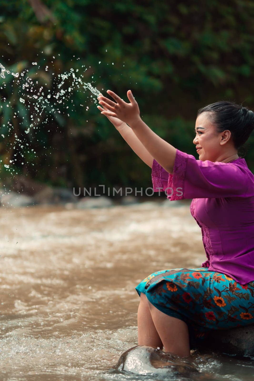 Happy Asian woman playing with the water near the river while sitting on the rock and wearing a purple dress inside the village