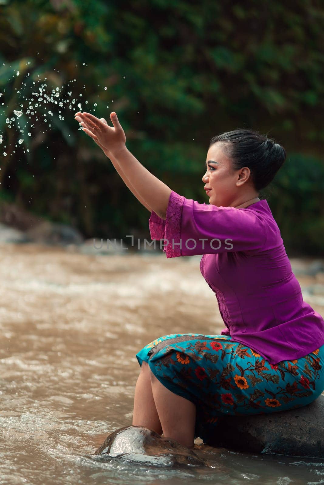 Happy Asian woman playing with the water near the river while sitting on the rock and wearing a purple dress inside the village