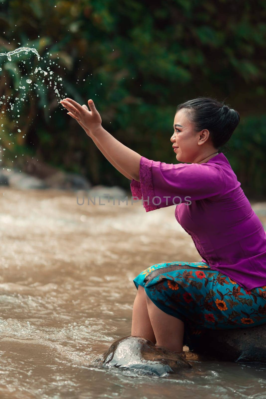 Happy Asian woman playing with the water near the river while sitting on the rock and wearing a purple dress inside the village