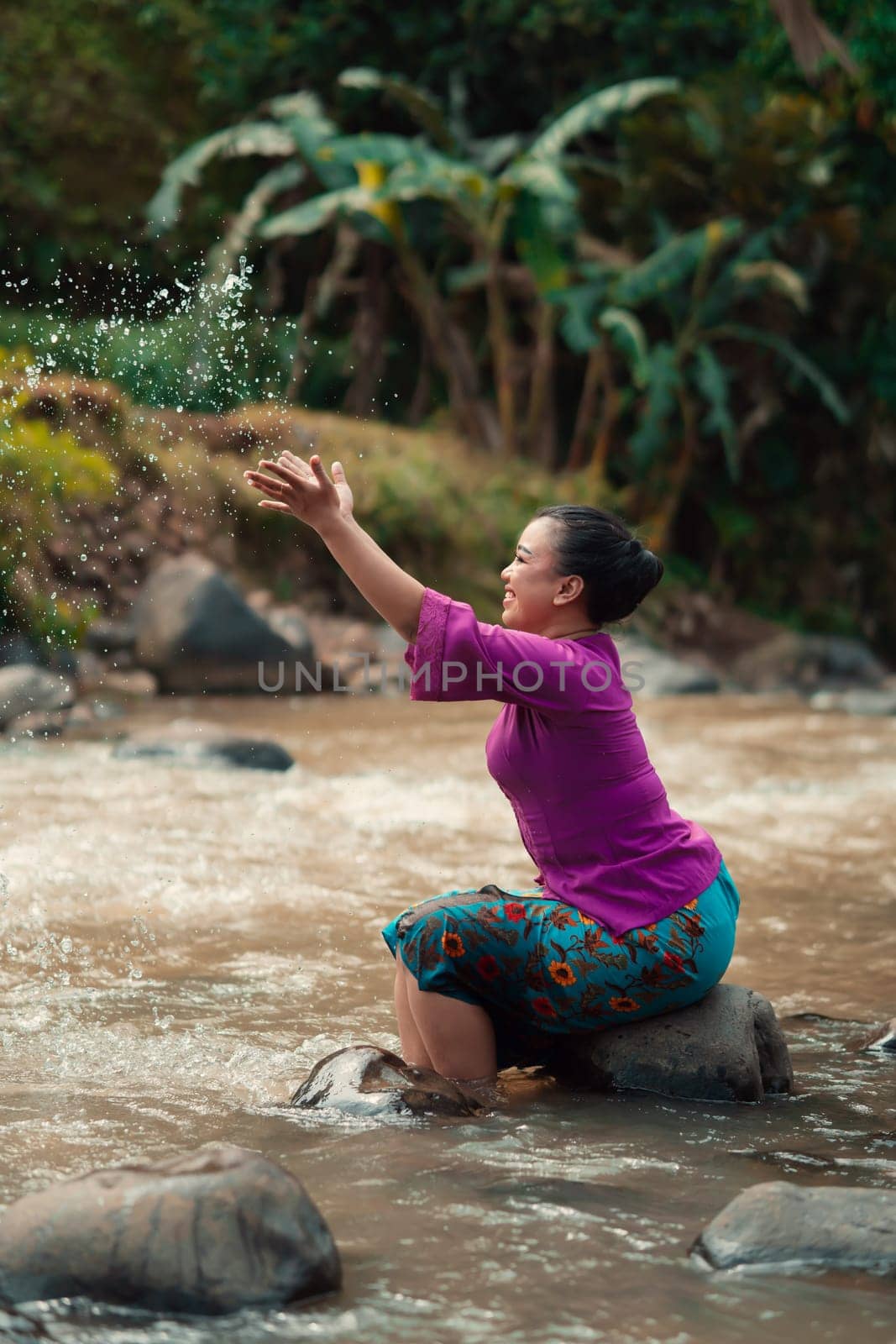 Happy Asian woman playing with the water near the river while sitting on the rock and wearing a purple dress inside the village