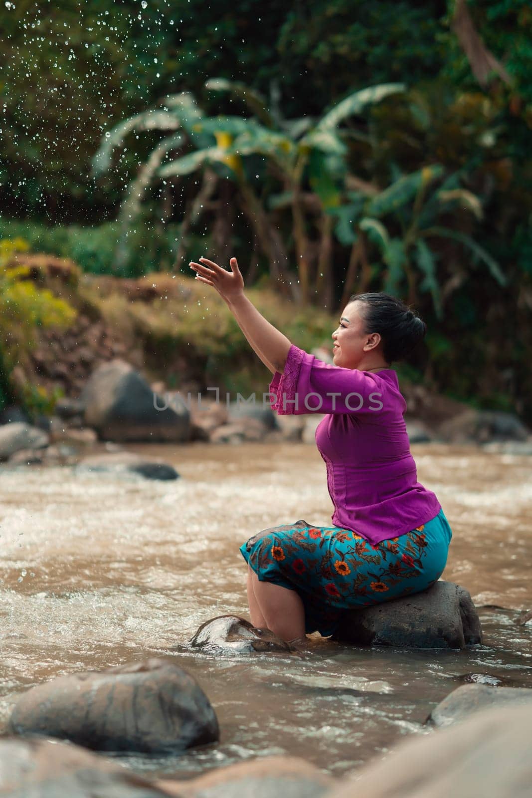 Indonesian woman sitting on a small rock and playing with the water while her dress got wet from the river near the rice field