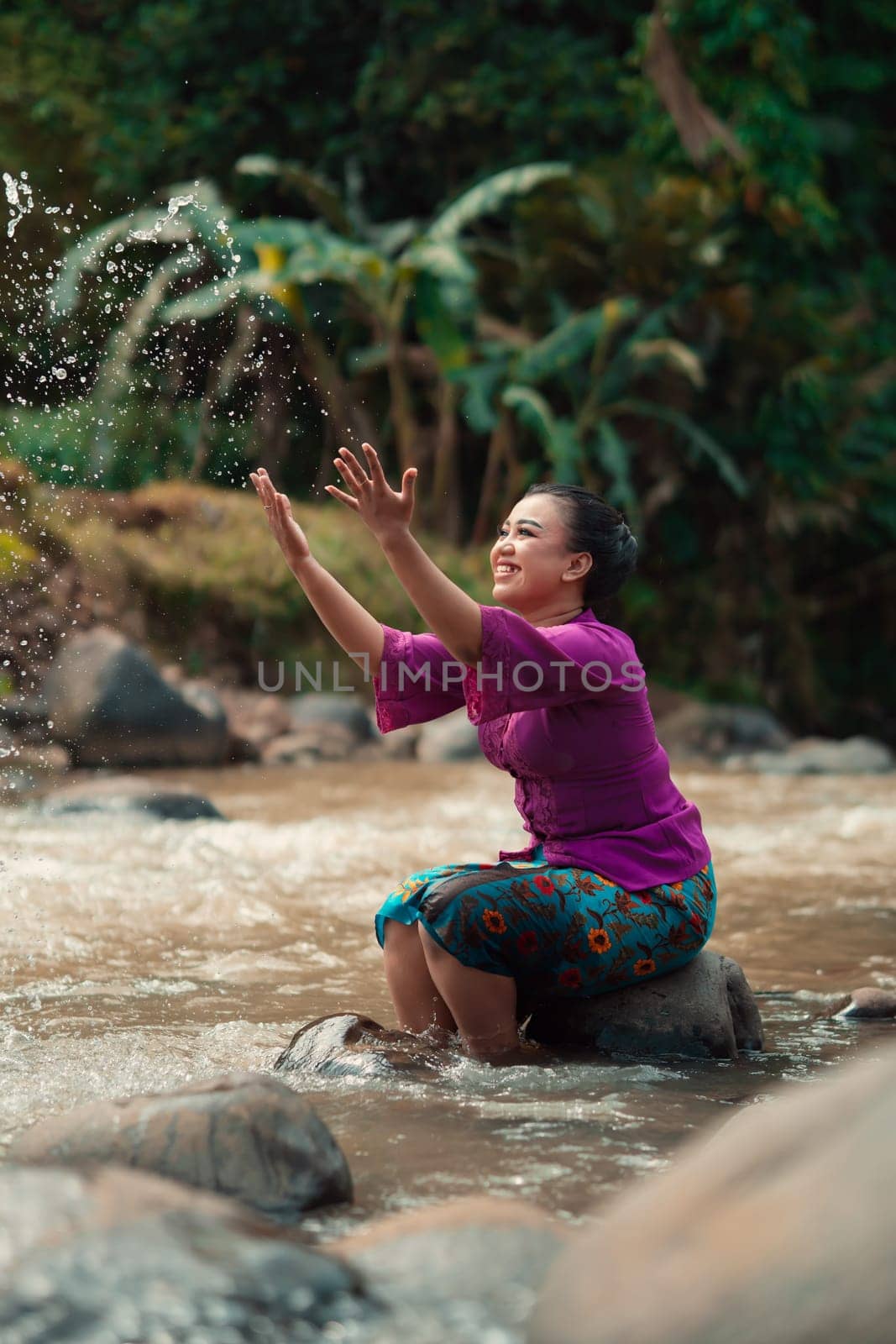 Indonesian woman sitting on a small rock and playing with the water while her dress got wet from the river near the rice field