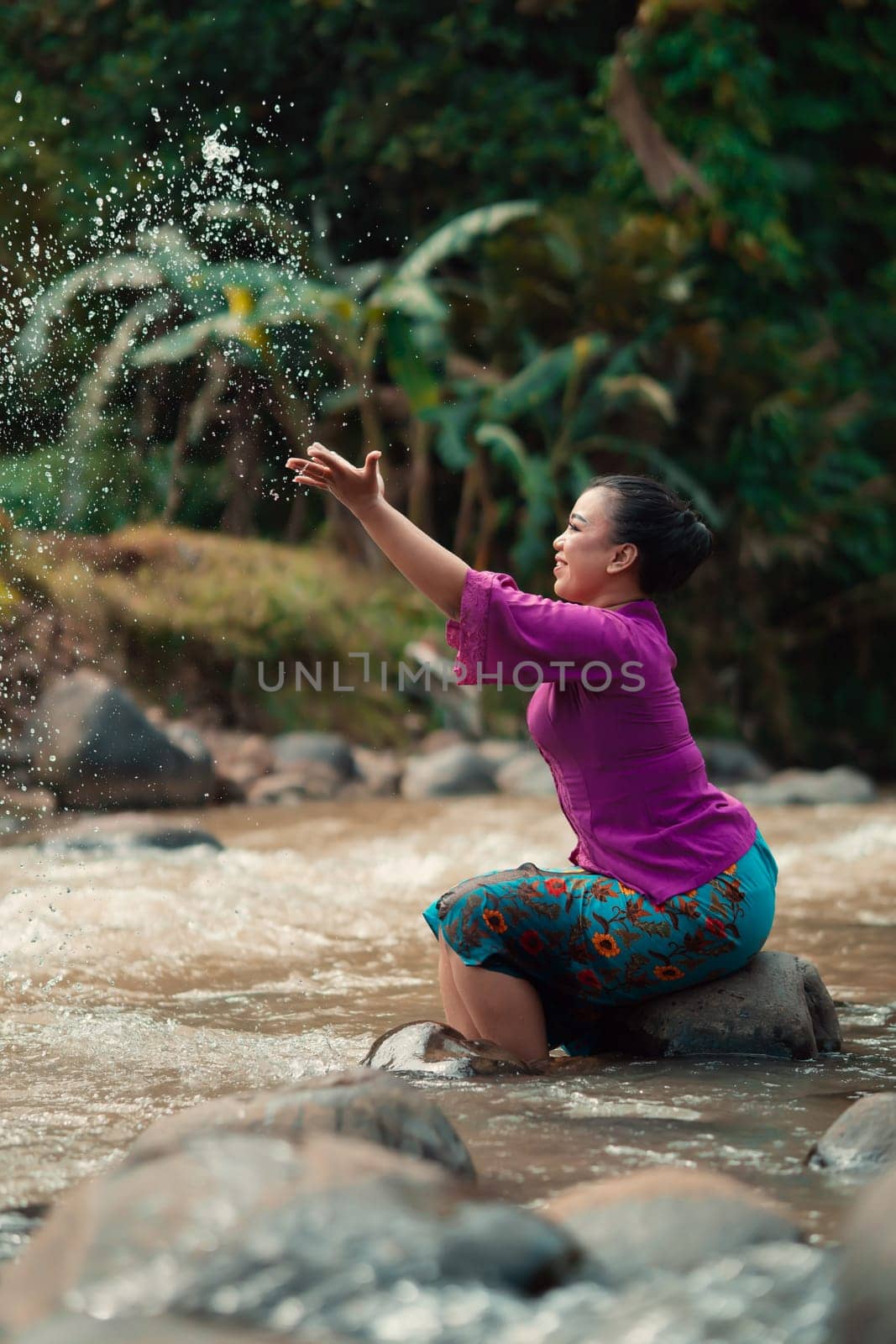 Indonesian woman sitting on a small rock and playing with the water while her dress got wet from the river near the rice field