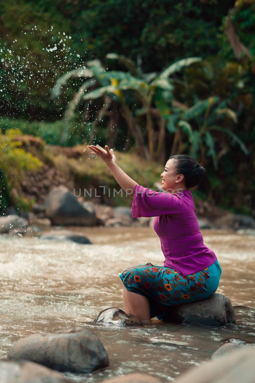 Indonesian woman sitting on a small rock and playing with the water while her dress got wet from the river near the rice field