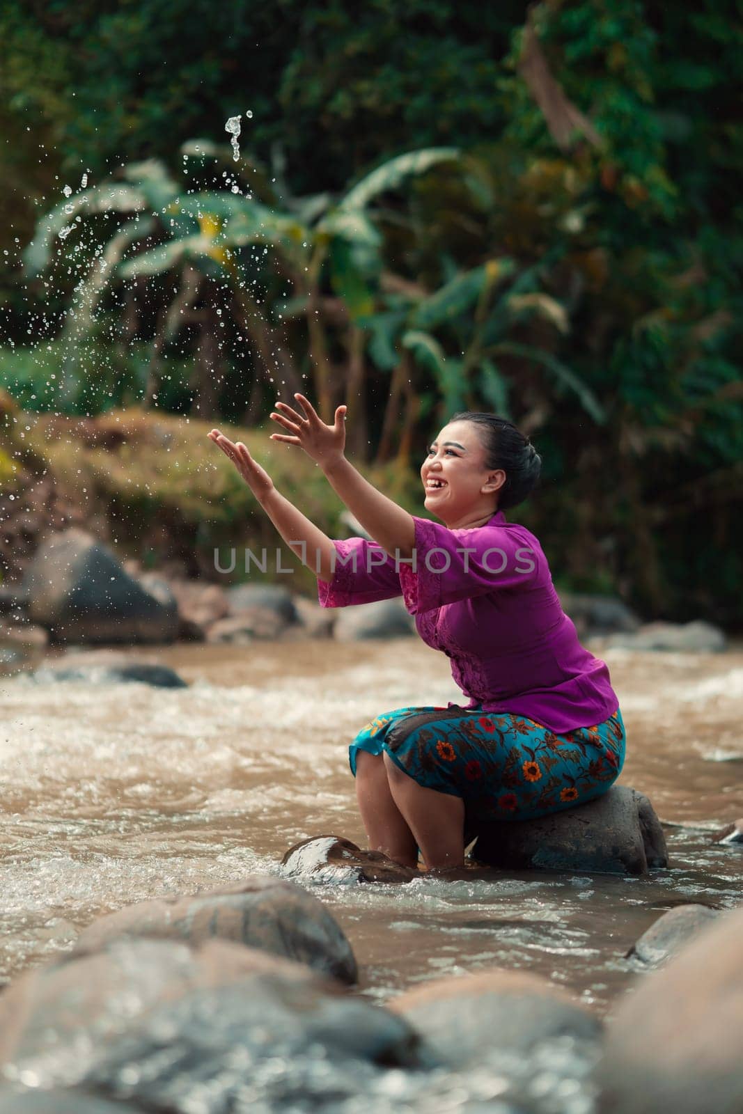Indonesian woman sitting on a small rock and playing with the water while her dress got wet from the river near the rice field