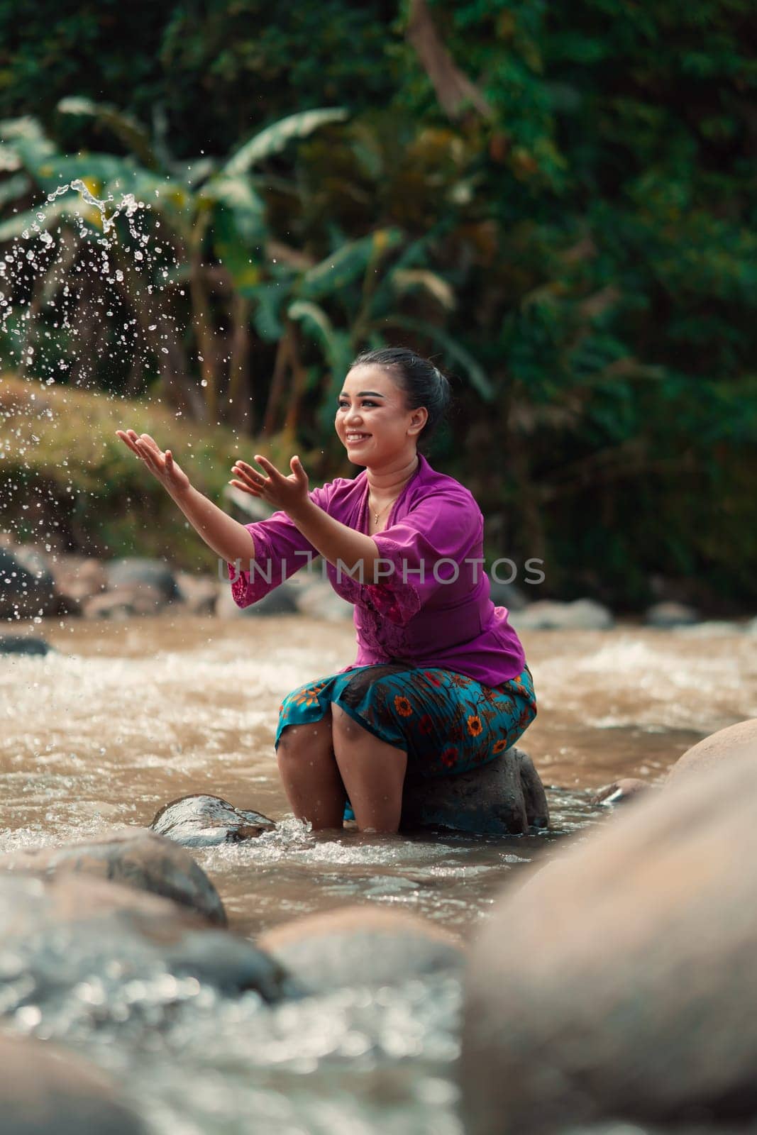 Asian woman playing with dirty water from a dirty river while wearing a purple dress and green skirt near the river