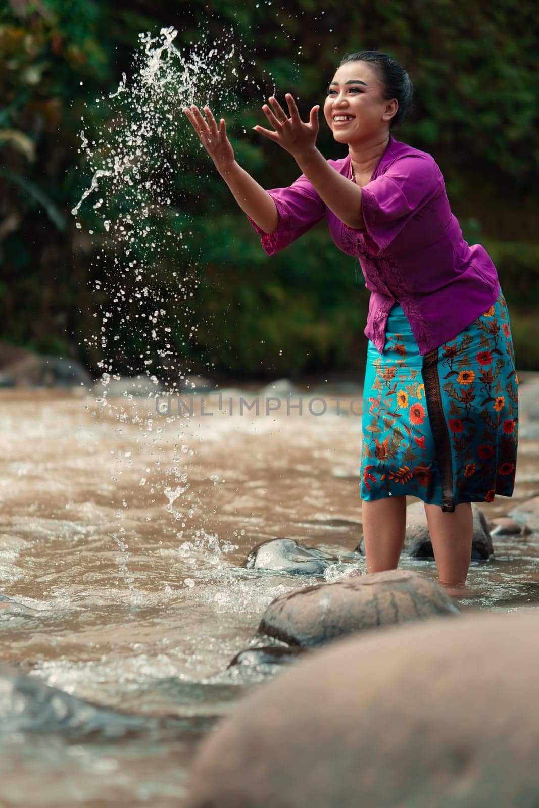 A beautiful Asian woman washing her hand and playing with the water while standing near the river in a traditional purple dress and green skirt in the village