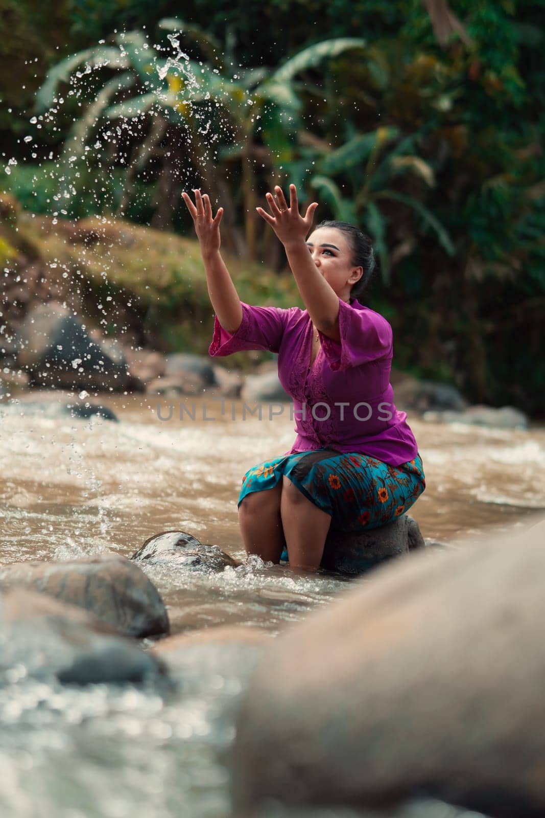 Asian woman playing with dirty water from a dirty river while wearing a purple dress and green skirt near the river