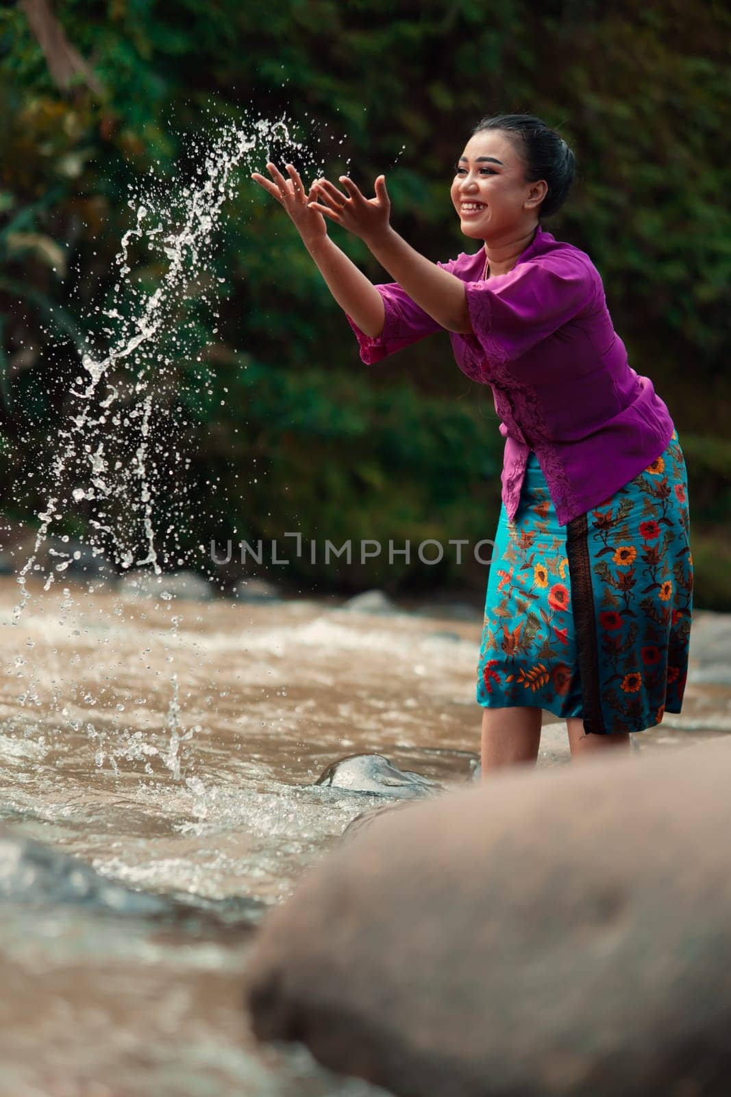 A beautiful Asian woman washing her hand and playing with the water while standing near the river in a traditional purple dress and green skirt in the village