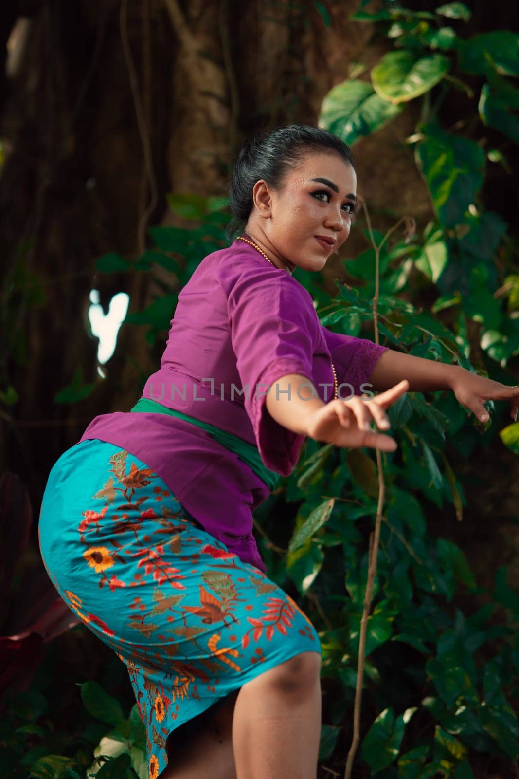 Sexy Sundanese woman with purple dress dancing in front of the big tree and the green leaves in the background during the daylight