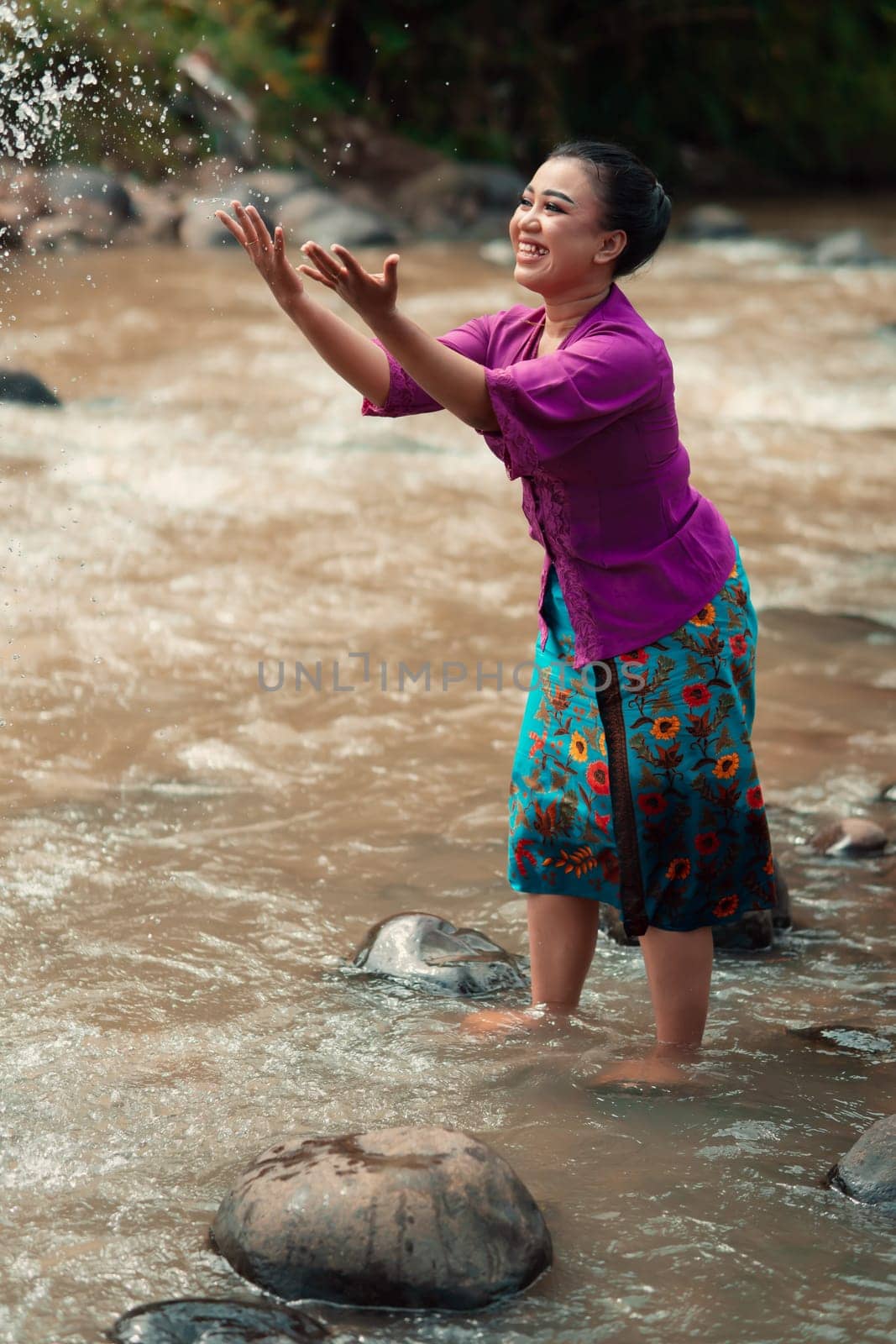 A beautiful Asian woman washing her hand and playing with the water while standing near the river in a traditional purple dress and green skirt in the village