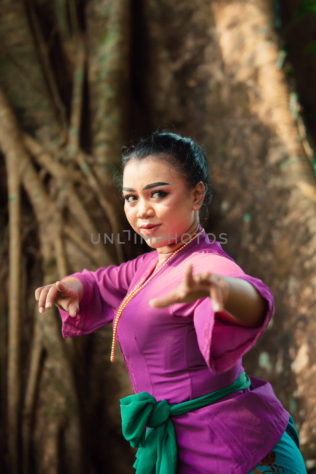 Sexy Asian woman dancing pose while wearing a beautiful purple dress and green belt on her body in front of the big brown tree inside the forest