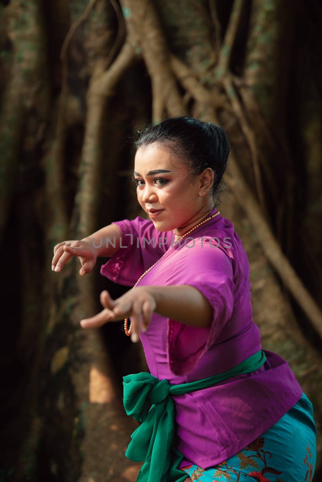 Sexy Asian woman dancing pose while wearing a beautiful purple dress and green belt on her body in front of the big brown tree inside the forest