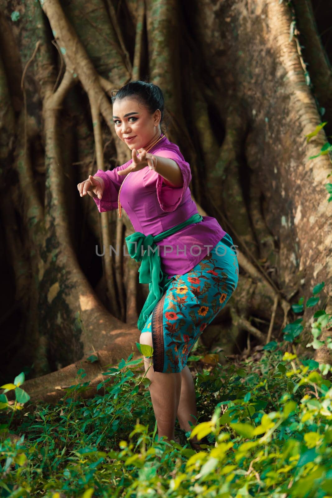 Sexy Asian woman dancing pose while wearing a beautiful purple dress and green belt on her body in front of the big brown tree inside the forest