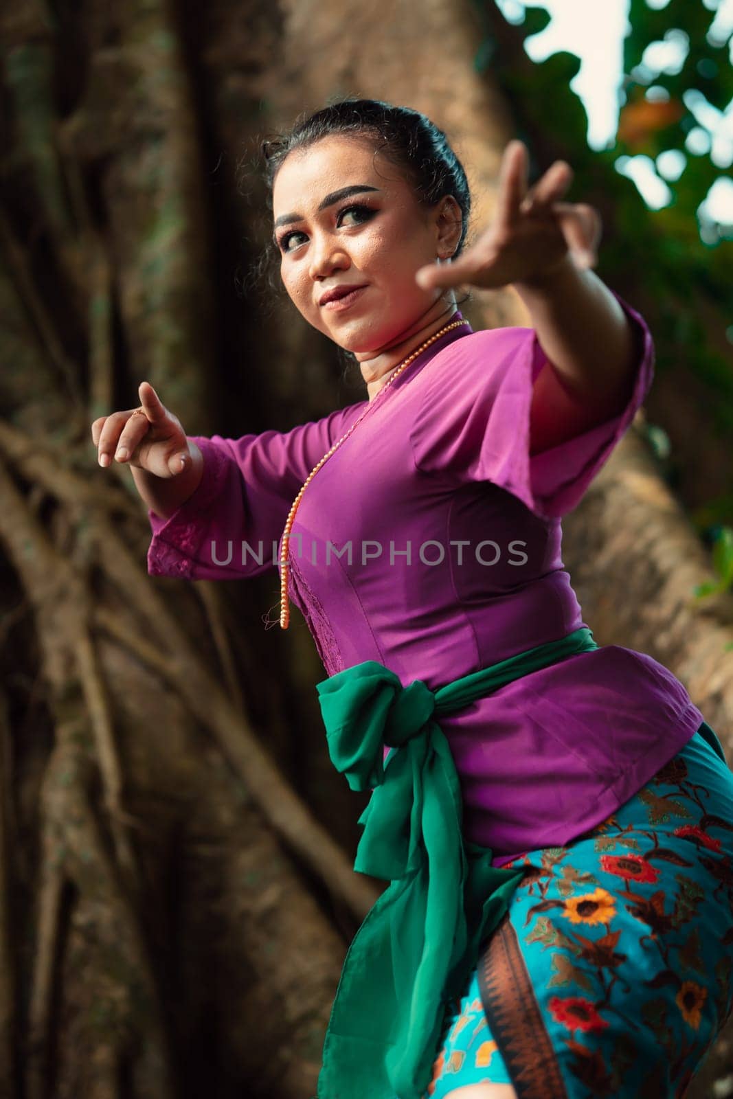 Sexy Asian woman dancing pose while wearing a beautiful purple dress and green belt on her body in front of the big brown tree inside the forest