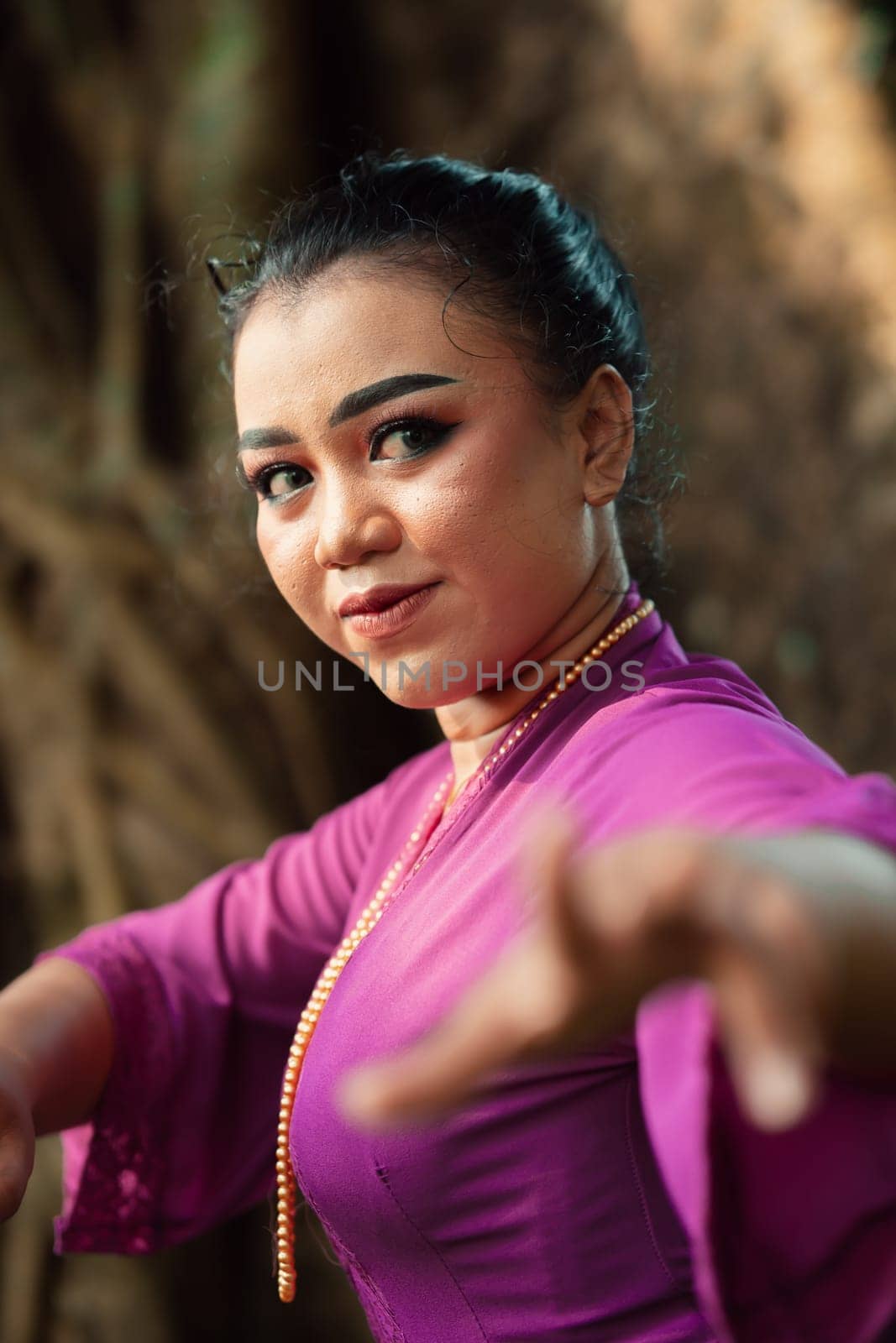 The face of a Javanese woman with makeup and a traditional purple dress called kebaya while dancing and posing with jewelry on her neck inside the jungle
