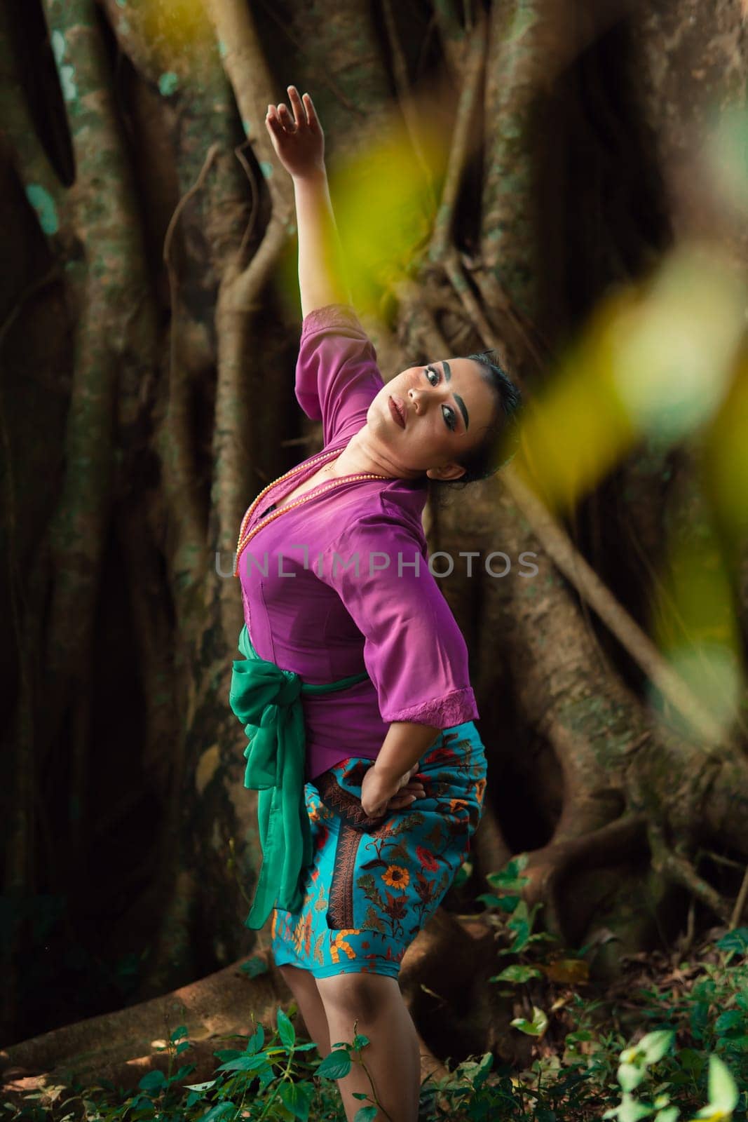 Asian woman takes a dancing pose in the jungle while wearing a purple dress and a golden necklace with a beautiful smile on her face
