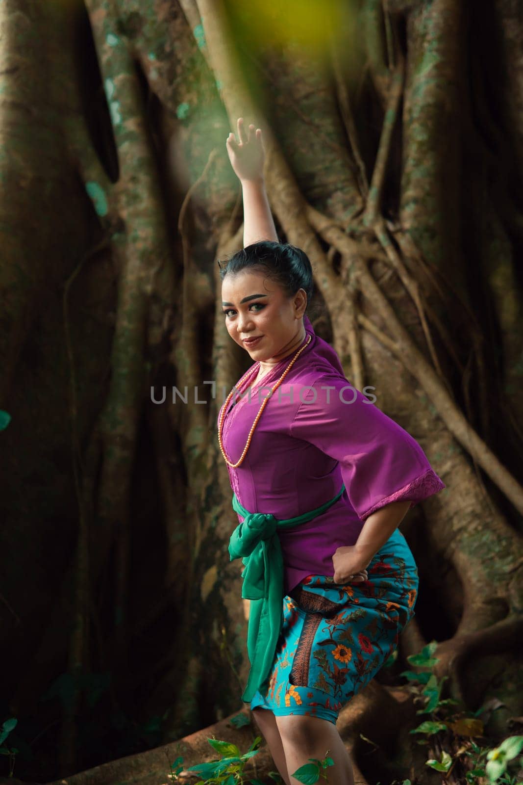 Asian woman takes a dancing pose in the jungle while wearing a purple dress and a golden necklace with a beautiful smile on her face